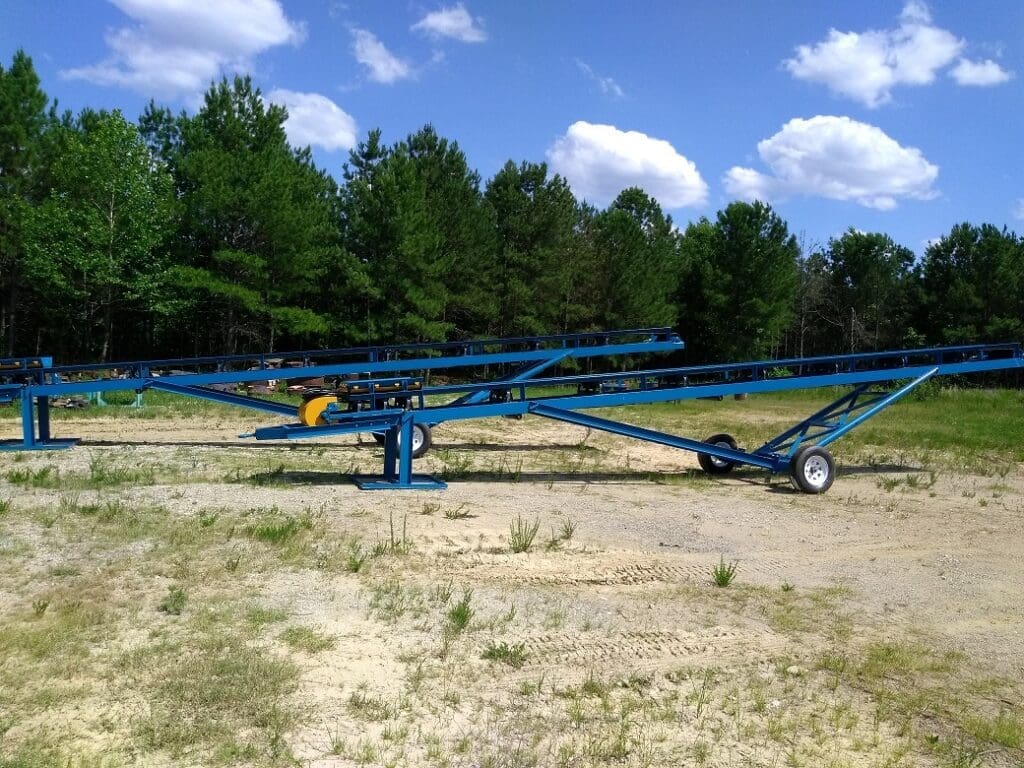 A blue metal conveyor system stands idle on a grassy, sandy surface, surrounded by trees under a clear, partly cloudy sky.